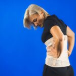 Woman in pain from back injury wearing lumbar brace corset on a blue background, copy space